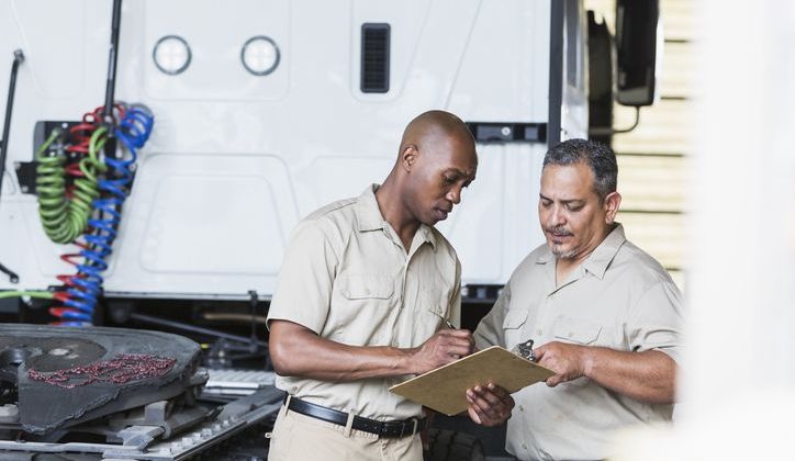 Two men next to a semi-truck in garage looking at a clipboard.