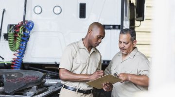 Two men next to a semi-truck in garage looking at a clipboard.