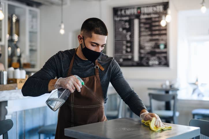Young man barista with face mask and gloves standing in coffee shop, disinfecting table
